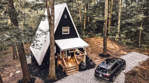 A cozy A-frame cabin surrounded by trees, with a gravel driveway and a parked car nearby.