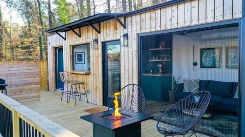 A cozy outdoor deck with modern seating, a fire pit, and wooden walls, surrounded by trees.