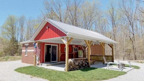 A small red cabin with a metal roof, surrounded by trees and gravel, featuring a porch and outdoor seating.
