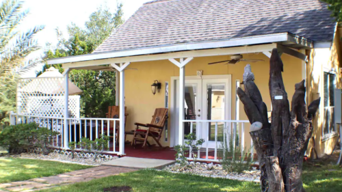 A cozy porch with rocking chairs, surrounded by greenery and a tree, in front of a charming yellow house.