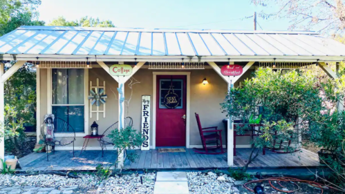 Charming cottage with a red door, porch, and signs welcoming friends, surrounded by greenery and gravel path.