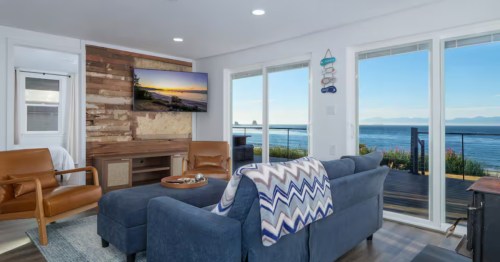 Cozy living room at Anchors Aweigh in Sekiu, Washington, with ocean views, blue sofa, leather chairs, and sliding doors to a waterfront deck.