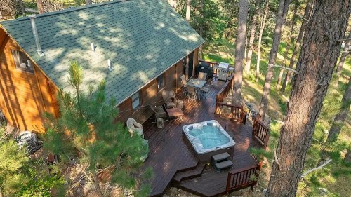 Aerial view of a wooden cabin surrounded by trees, featuring a deck with a hot tub and outdoor seating.