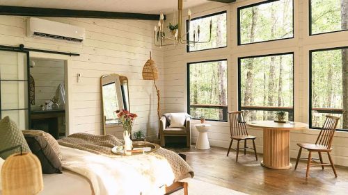 Cozy bedroom with large windows, wooden furniture, and a serene forest view, featuring a bed, table, and chairs.