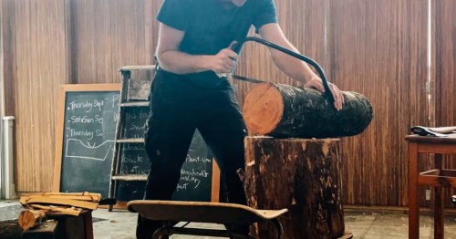 Woodworker using a hand saw to cut a log on a rustic chopping block in a sunlit studio with a chalkboard nearby.