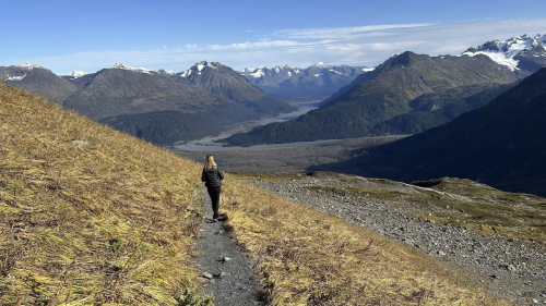 A person walks along a trail in a mountainous landscape with valleys and snow-capped peaks in the background.