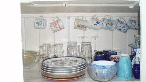 A wooden cupboard filled with various dishes, glasses, and hanging mugs in different colors and designs.