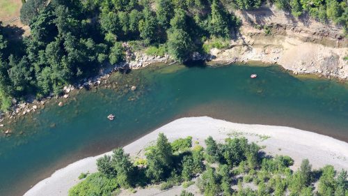 Aerial view of a winding river surrounded by lush greenery and sandy banks, with two small boats visible on the water.