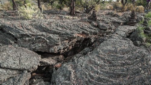 Lava rock formations with distinct flow patterns and a small stack of stones among sparse vegetation.