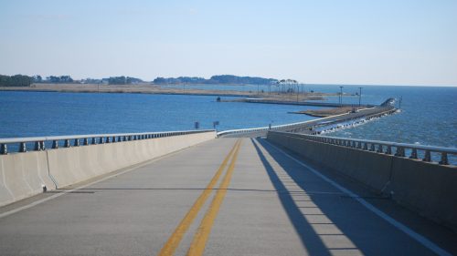 A wide road leads towards a bridge over water, with an island visible in the distance under a clear blue sky.