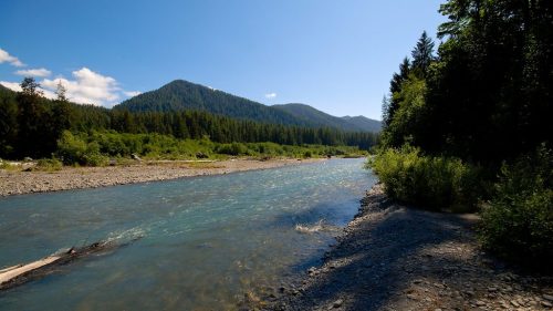 A serene river flows through a lush green landscape, surrounded by mountains under a clear blue sky.