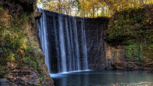 A serene waterfall cascading over rocks into a calm pool, surrounded by lush greenery and autumn foliage.