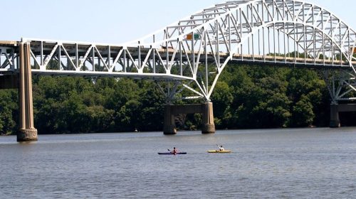Two kayakers paddle on a river beneath a large white bridge surrounded by green trees.