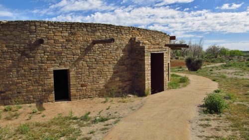 A stone building with two doorways, surrounded by a dirt path and sparse vegetation under a blue sky with clouds.