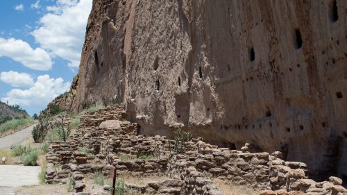 Ancient stone ruins beside a cliff, with small caves and a clear blue sky in the background.