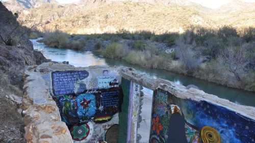 A colorful mural on a stone wall overlooks a river and mountains in a serene landscape.