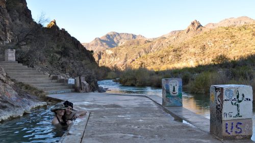 Two people relax in a natural hot spring by a river, surrounded by rocky hills and greenery under a clear sky.