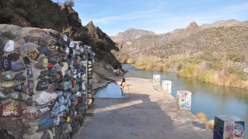 A scenic riverbank with colorful painted rocks and a person walking along the path, surrounded by mountains and greenery.