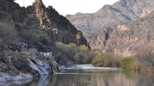 A serene river flows between rocky hills, with sparse vegetation and a palm tree along the bank under a cloudy sky.