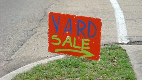 Bright orange yard sale sign with blue and green lettering, pointing right on a grassy area near a road.