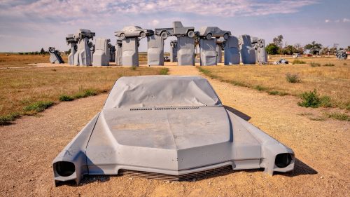 A large, gray car sculpture in the foreground with a backdrop of stacked car sculptures against a blue sky.