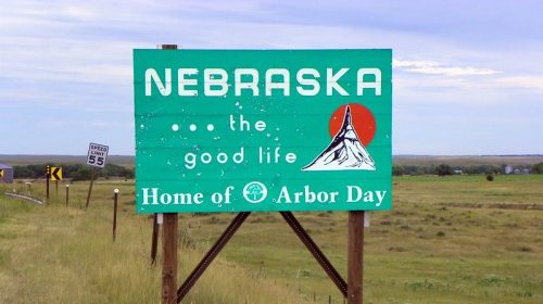 Green sign welcoming visitors to Nebraska, highlighting "the good life" and celebrating Arbor Day. Scenic landscape in background.