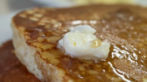 Close-up of fluffy pancakes topped with melting butter and syrup, with a plate of scrambled eggs in the background.