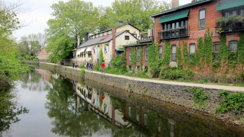 A serene canal lined with greenery and brick buildings, reflecting the scene in calm water, with people walking nearby.