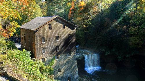 A historic wooden mill beside a waterfall, surrounded by colorful autumn foliage and a tranquil river.