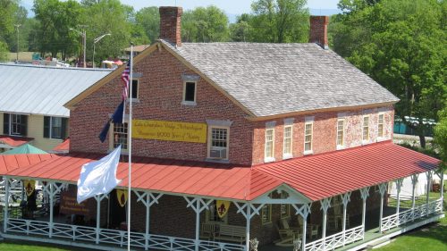 Historic brick building with a red roof, surrounded by green grass and trees, featuring a flagpole in front.