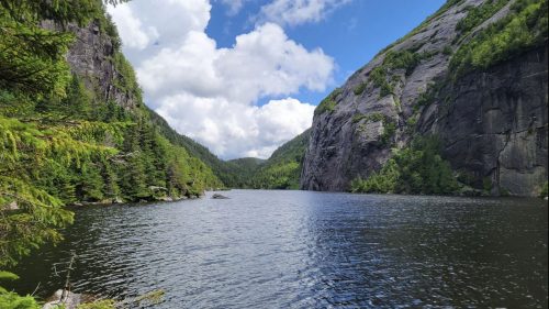 A serene lake surrounded by lush green trees and steep rocky cliffs under a partly cloudy sky.