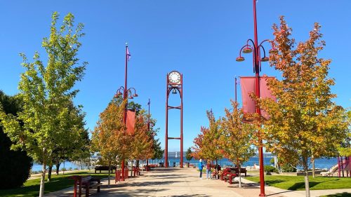 A sunny park pathway lined with trees and red banners, featuring a tall clock tower and views of the water.