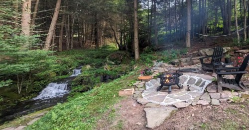 Outdoor fire pit on a stone patio next to a small forest waterfall with black lounge chairs.