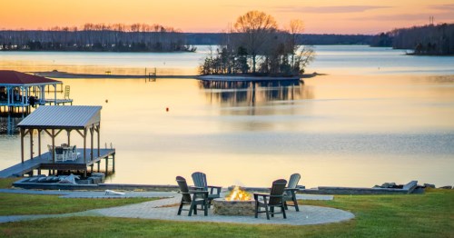 A circle of chairs around a firepit with a lake and dock in the background with a setting sun.