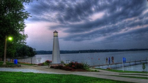 A lighthouse stands by a lake at dusk, with people walking along the shore under a cloudy sky.