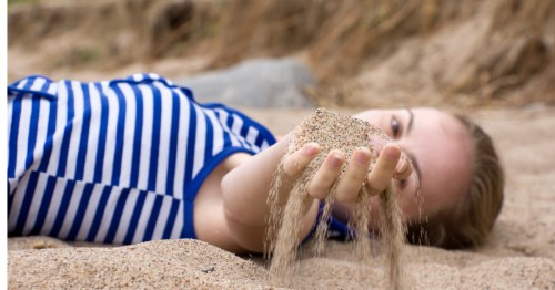 A woman wearing a blue and white striped shirt laying in the sand with her hand in front of her face, sand trickling through her fingers. She's at Sand Prairie Conservation Area, on a dune hike in Missouri.