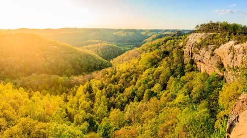A scenic view of a lush green valley at sunset, with rolling hills and a rocky cliff in the foreground.