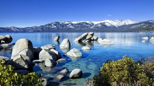 A serene lake scene with clear blue water, rocky formations, and snow-capped mountains under a bright blue sky.