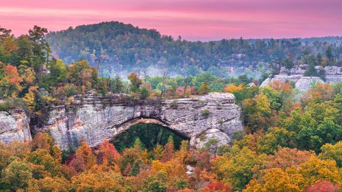 A scenic view of a natural stone arch surrounded by vibrant autumn foliage and misty hills at sunrise.