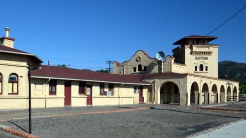 Historic train station with a yellow exterior, red accents, and a tower, set against a clear blue sky.