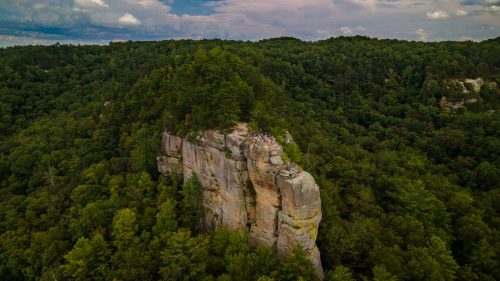 Aerial view of a rocky cliff surrounded by lush green trees under a partly cloudy sky.