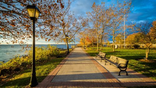 A scenic waterfront path lined with benches and trees, illuminated by a lamp under a partly cloudy sky.