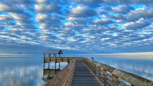 A wooden pier extends over calm water, leading to a small shelter under a sky filled with fluffy clouds.