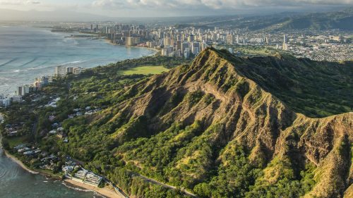 Aerial view of Diamond Head crater with Honolulu skyline and coastline in the background under a blue sky.