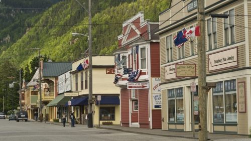 A quaint street lined with colorful buildings, shops, and flags, set against a backdrop of green mountains.