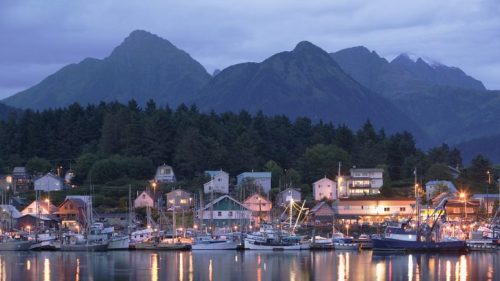A serene harbor at dusk, with boats docked and mountains in the background, illuminated by soft lights from nearby buildings.