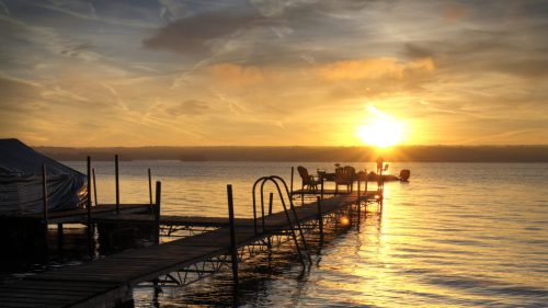 A serene sunset over a calm lake, with a wooden dock extending into the water and silhouettes of chairs in the distance.