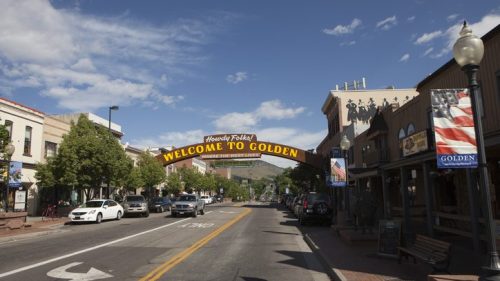A street view of Golden, Colorado, featuring a welcoming arch and shops under a blue sky with scattered clouds.
