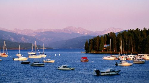 A serene lake scene with various boats anchored, surrounded by mountains and trees under a soft pink sky.