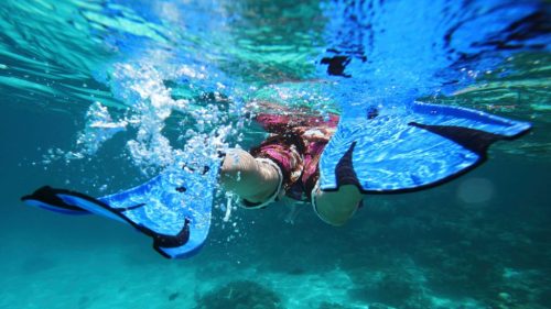 A person snorkeling underwater, wearing fins, with bubbles and vibrant blue water surrounding them.